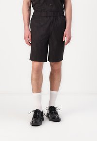 Shorts noirs avec des rayures verticales, ceinture élastique, poches latérales, associés à des chaussettes blanches mi-mollet et des chaussures noires à lacets.