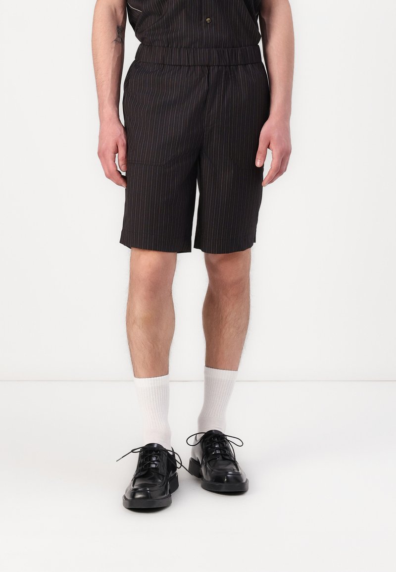 Shorts noirs avec des rayures verticales, ceinture élastique, poches latérales, associés à des chaussettes blanches mi-mollet et des chaussures noires à lacets.