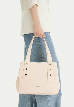 Osoba držící béžovou koženou kabelku se širokými popruhy a kovovými nýty, oblečená v lehkém saku a modrých džínách, na jednobarevném pozadí.