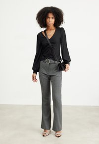 Black long-sleeved top with gathered front detail, grey patterned wide-leg trousers, silver belt buckle, and black handbag. Black heeled sandals.