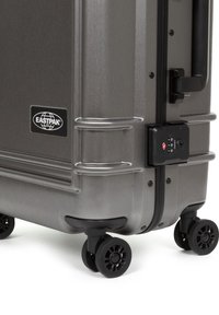 Gray hard-shell suitcase by Eastpak with a textured surface, reinforced corners, side handle, combination lock, and four black wheels.