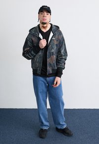 Camouflage zip-up jacket with black accents, black t-shirt, loose blue jeans, and black sneakers. Black cap with logo.