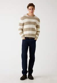 Beige és fehér csíkos kötött pulóver bordázott textúrával, sötétkék farmernadrággal és sötétbarna cipővel párosítva. A modell a zsebébe dugott kézzel áll.