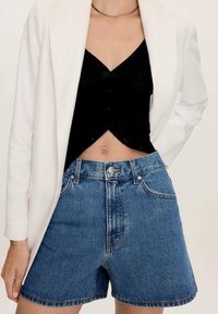 Shorts de mezclilla de cintura alta, lavado azul medio y corte relajado, combinados con un top corto negro y una americana blanca. Collar de cadena plateada.