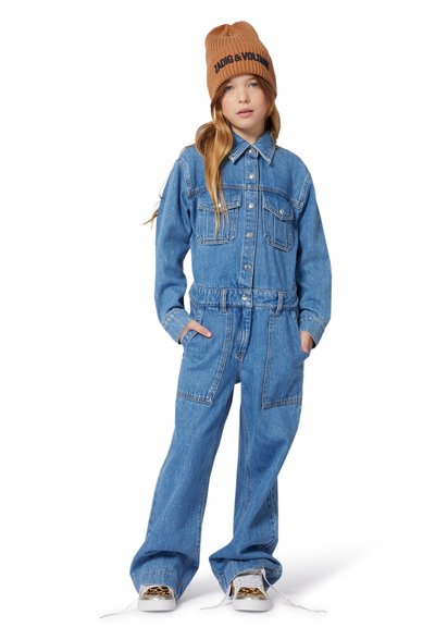 Meisje met een bruine muts, blauwe spijkerjack en broek, staand met handen in de zakken, witte sneakers met leopardenprint op de neus.