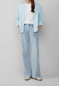 Jasnoniebieska koszula zapinana na guziki nałożona na białą koszulkę, w połączeniu z luźnymi jasnoniebieskimi dżinsami i beżowym obuwiem.