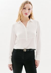 Blusa blanca de manga larga con cuello y textura de rayas verticales, combinada con jeans negros y un cinturón con detalles en dorado.