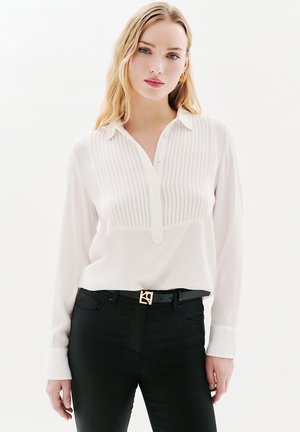 Blusa bianca a maniche lunghe con colletto e texture a righe verticali, abbinata a jeans neri e una cintura con accenti dorati.