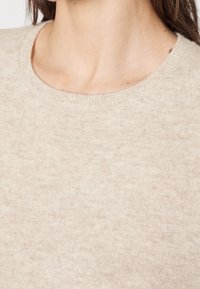 Beige strikket sweater med rund hals og ribbede kanter. Blød tekstur med et subtilt mønster. Nærbillede, der viser halsudskæringen og stofdetaljerne.