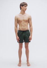 Πράσινα μαγιό βερμούδα με κορδόνι στη μέση. Ελαφρύ ύφασμα. Χωρίς ορατά σχέδια ή διακοσμήσεις. Το μοντέλο στέκεται ξυπόλητο σε απλό φόντο.