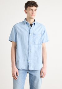 Camisa de mezclilla de manga corta en azul claro con cuello, cierre de botones en el frente y bolsillo en el pecho izquierdo. La tela parece suave con una textura ligeramente desgastada.
