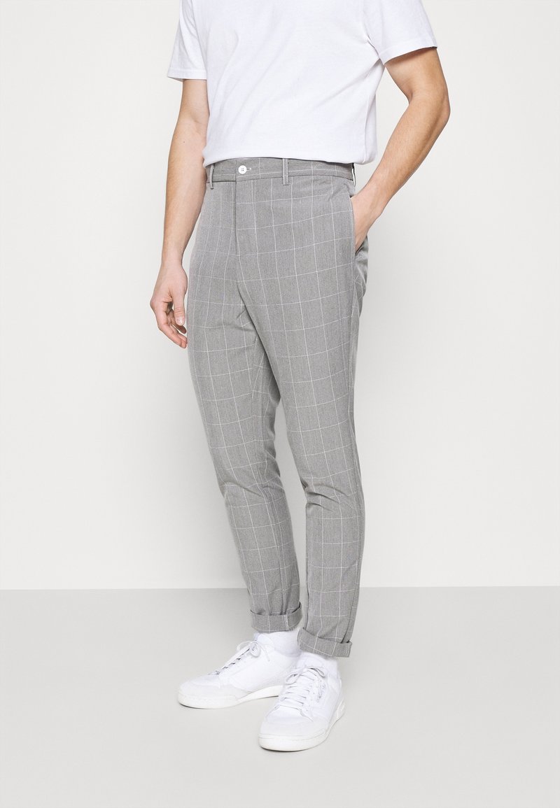 Muž ve světle šedých kostkovaných slim-fit kalhotách, bílých teniskách a bílém tričku, stojící s jednou rukou v kapse.