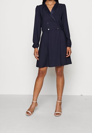 Femme portant une robe portefeuille bleu marine à manches longues avec des boutons croisés et des talons blancs ouverts avec brides à la cheville.