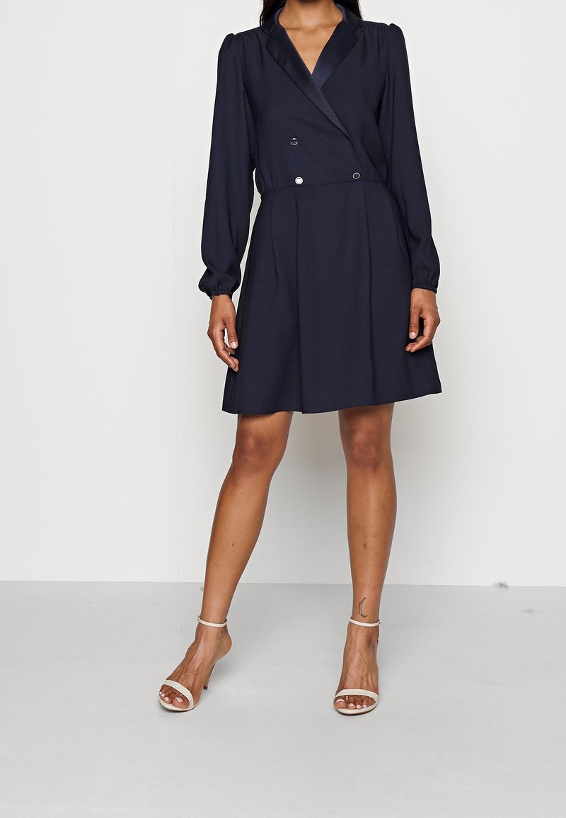 Femme portant une robe portefeuille bleu marine à manches longues avec des boutons croisés et des talons blancs ouverts avec brides à la cheville.