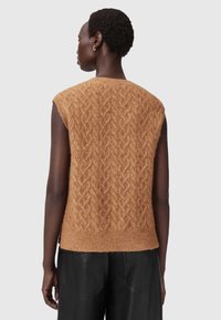 Brun strikket vest med kabelmønster, ærmeløs design, blød tekstur og ribbet kant. Båret med sorte bukser.