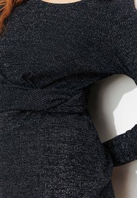 Robe en maille noire avec des détails en fil métallique, présentant un design avant torsadé et des manches longues. Texture lisse et silhouette ajustée.