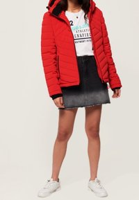 Czerwona pikowana kurtka z kapturem, czarne akcenty, noszona na białej koszulce z grafiką i czarnej d�żinsowej spódnicy, w połączeniu z białymi trampkami.