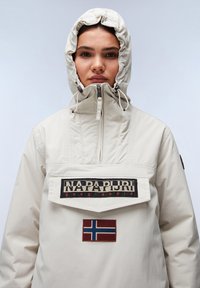 Giacca pullover beige chiaro con colletto alto, cappuccio regolabile e patch con logo. Presenta una patch con la bandiera norvegese e un tessuto strutturato.