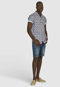 Korte mouwen blauwe blouse met bloemenpatroon, denim shorts en beige instapschoenen. Heeft opgezoete mouwen en een rechte snit.