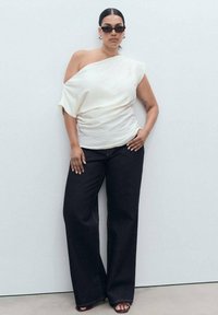 Personne portant un pantalon large noir, un haut blanc dénudé aux épaules, des lunettes de soleil sombres, se tenant contre un mur blanc uni avec une main sur la hanche.