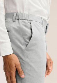 Helle graue Anzughose mit glatter Textur, ausgestattet mit Falten, seitlichen Taschen und einem maßgeschneiderten Schnitt, zusammen mit einem weißen, strukturierten Hemd präsentiert.