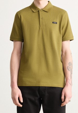 Homme portant un polo à manches courtes vert olive avec un patch logo Calvin Klein noir et un pantalon noir, montrant des tatouages sur l'avant-bras droit.