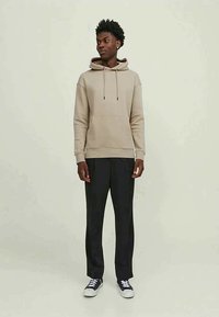 Giovane in piedi, con una felpa beige, pantaloni neri e sneakers bianco e nere, su uno sfondo chiaro e uniforme.