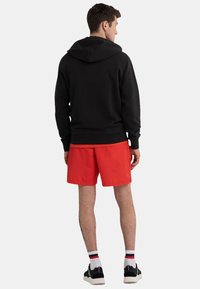 Sweat à capuche noir avec une poche kangourou, associé à un short rouge. Le modèle porte des chaussettes blanches et rouges rayées et des baskets noires.