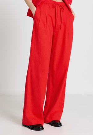 Pantalon rouge à jambes larges avec plis devant et taille à cordon, porté avec des chaussures noires et les mains dans les poches.