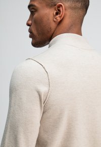 Beige gebreide pullover met een hoge kraag, met een gestructureerde afwerking en contrasterende stiksels langs de schoudernaad.