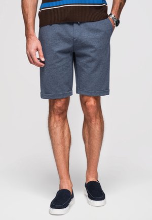 Mann trägt blaue gemusterte Shorts, dunkle Slipper mit weißen Sohlen und einen braunen Pullover mit blauen und weißen Streifen, steht mit entspannten Händen.