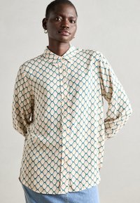 Button-up shirt in cream with a geometric teal pattern, featuring a classic collar, long sleeves, and a relaxed fit. Soft fabric texture.