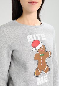 Szary sweatshirt z grafiką piernikowego ludka w czerwonej czapce Mikołaja oraz napisem "BITE ME". Materiał ma miękką fakturę.