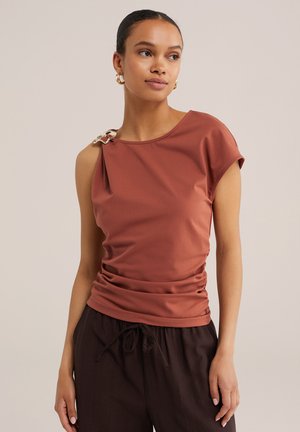 Femme portant un haut sans manches couleur rouille avec une boucle décorative sur une épaule et un pantalon noir à cordon, debout devant un fond neutre.