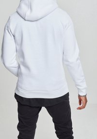 Witte hoodie met een capuchon met trekkoord, geribbelde manchetten en zoom. Gecombineerd met zwarte broek, casual achteraanzicht op een lichte achtergrond.