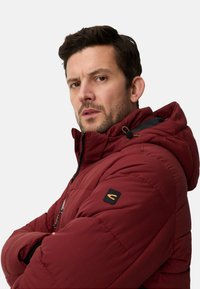 Czerwona pikowana kurtka z kapturem, zapinana na zamek, z czarnym akcentem na ramieniu oraz fakturowaną tkaniną z detalami szwów.