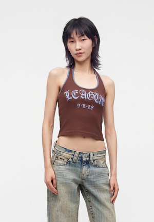 Person med glat sort hår har en brun crop top med teksten "LEAGUE 9-1-98" og falmede blå jeans på og står mod en ensfarvet baggrund.