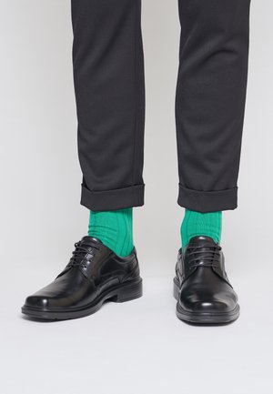 Czarne eleganckie buty noszone z podwiniętymi czarnymi spodniami i jaskrawozielonymi prążkowanymi skarpetami na jednolitym jasnym tle.