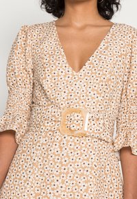 Donna che indossa un vestito beige con stampa floreale bianca, maniche a palloncino, scollatura a V profonda e una cintura coordinata con una grande fibbia quadrata.