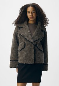 Cappotto in finta pelliccia di pecora marrone con colletto a punta, grandi tasche frontali e rifiniture nere. Abbinato a una gonna scura per un look stratificato.