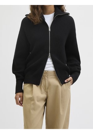 Femme portant un pull noir zippé sur une chemise blanche et un pantalon plissé beige, une main dans la poche et des cheveux bouclés visibles.