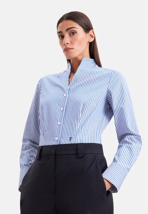 Donna con capelli lisci e scuri che indossa una camicia a righe verticali blu e bianche abbottonata, infilata in pantaloni neri a vita alta, mani nelle tasche.