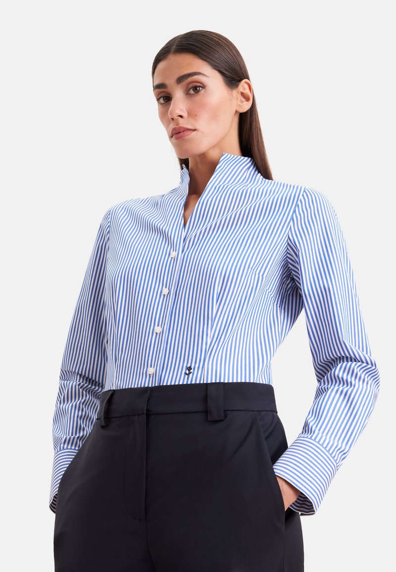 Femme aux cheveux lisses et foncés portant une chemise à rayures verticales bleues et blanches, boutonnée et rentrée dans un pantalon noir taille haute, les mains dans les poches.