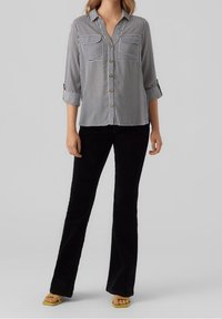 Camicia a righe con due tasche sul petto, maniche arrotolate; abbinata a pantaloni neri svasati e sandali gialli.