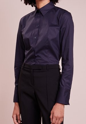 Vrouw die een aansluitend, donkerblauw overhemd met lange mouwen draagt, ingestopt in een zwarte hoge taille broek, voor een neutrale achtergrond.