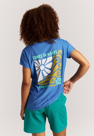 Femme aux cheveux bouclés portant une chemise bleue avec des motifs de soleil, vague et fleur, et le texte « Comes in Waves » dans le dos, associée à un short vert.
