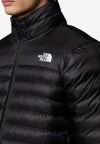Črna puhovka z visokim ovratnikom, prešita tekstura in belim logotipom na levi prsni strani. Ima sprednji zadrgo in gladek material.