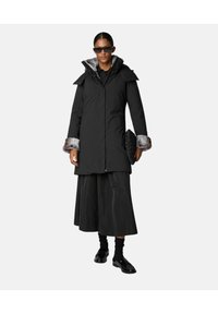 Sort knælang parka med faux fur manchetter og krave, parret med en lang sort nederdel og tilbehør. Har en frontlynlås og lommer.