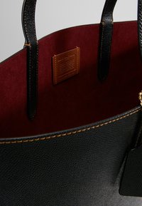 Czarna skórzana torba tote o gładkiej fakturze, bordowa zamszowa podszewka, brązowa metka z logo oraz kontrastowe złote przeszycia wzdłuż krawędzi.