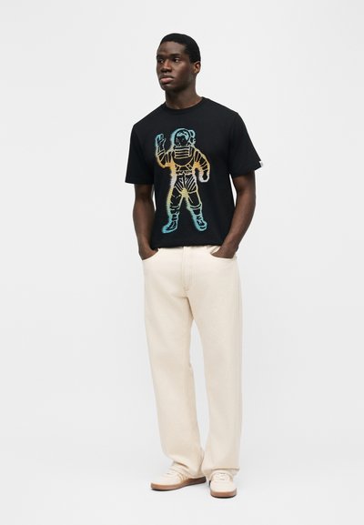 Άνδρας που φοράει μαύρο μπλουζάκι με πολύχρωμο γραφικό αστροναύτη, ευρύχωρο κρεμ παντελόνι και λευκά αθλητικά παπούτσια, μπροστά σε απλό φωτεινό φόντο.